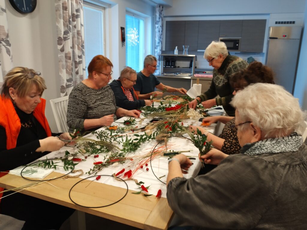 Kuvassa senioritalon asukkaat askartelevat joulukransseja tuutorin kanssa.